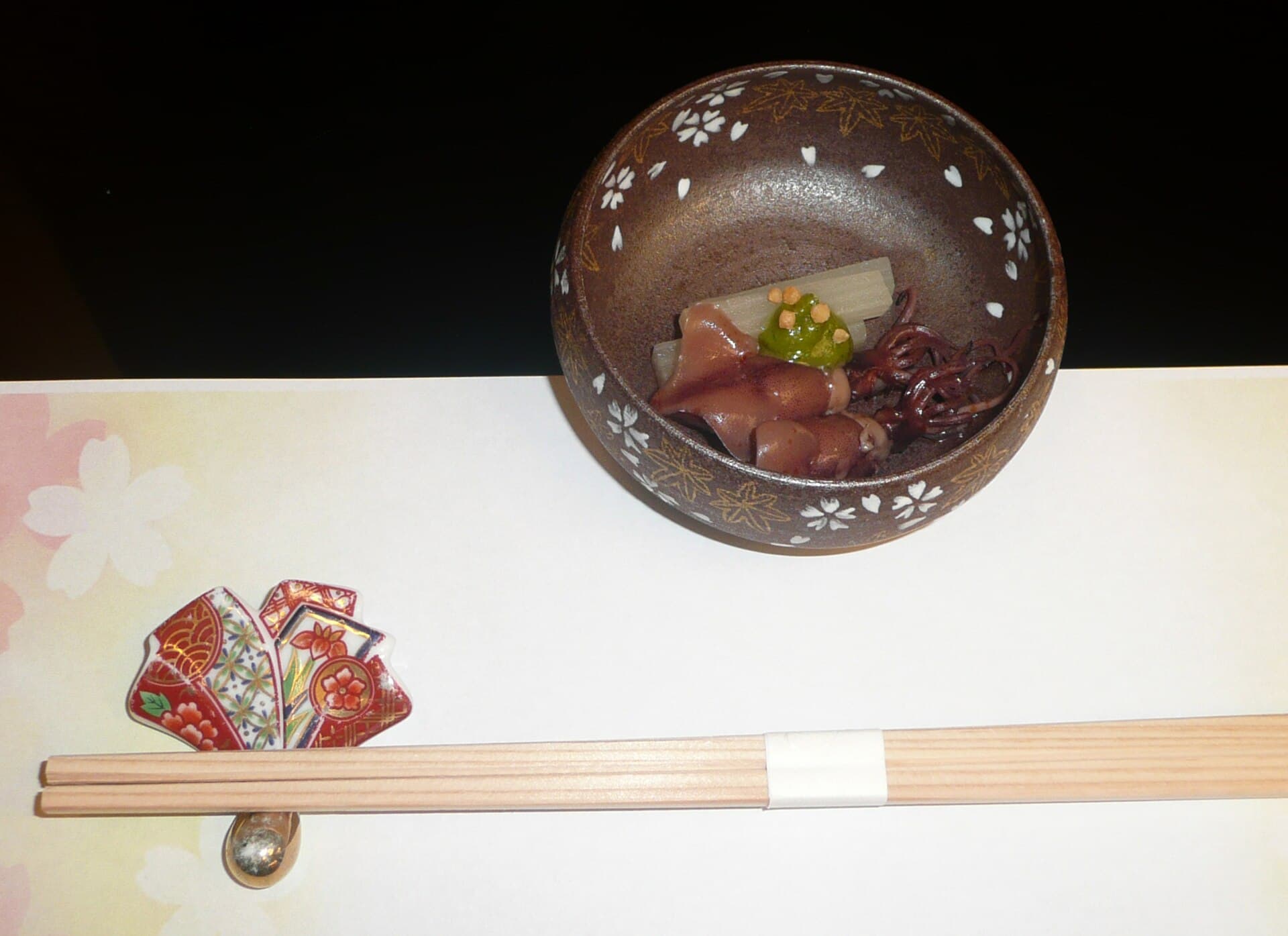 A brown ceramic bowl with maple-leaf and blossom patterns holds a small squid dish, beside pale wood chopsticks on a rest.