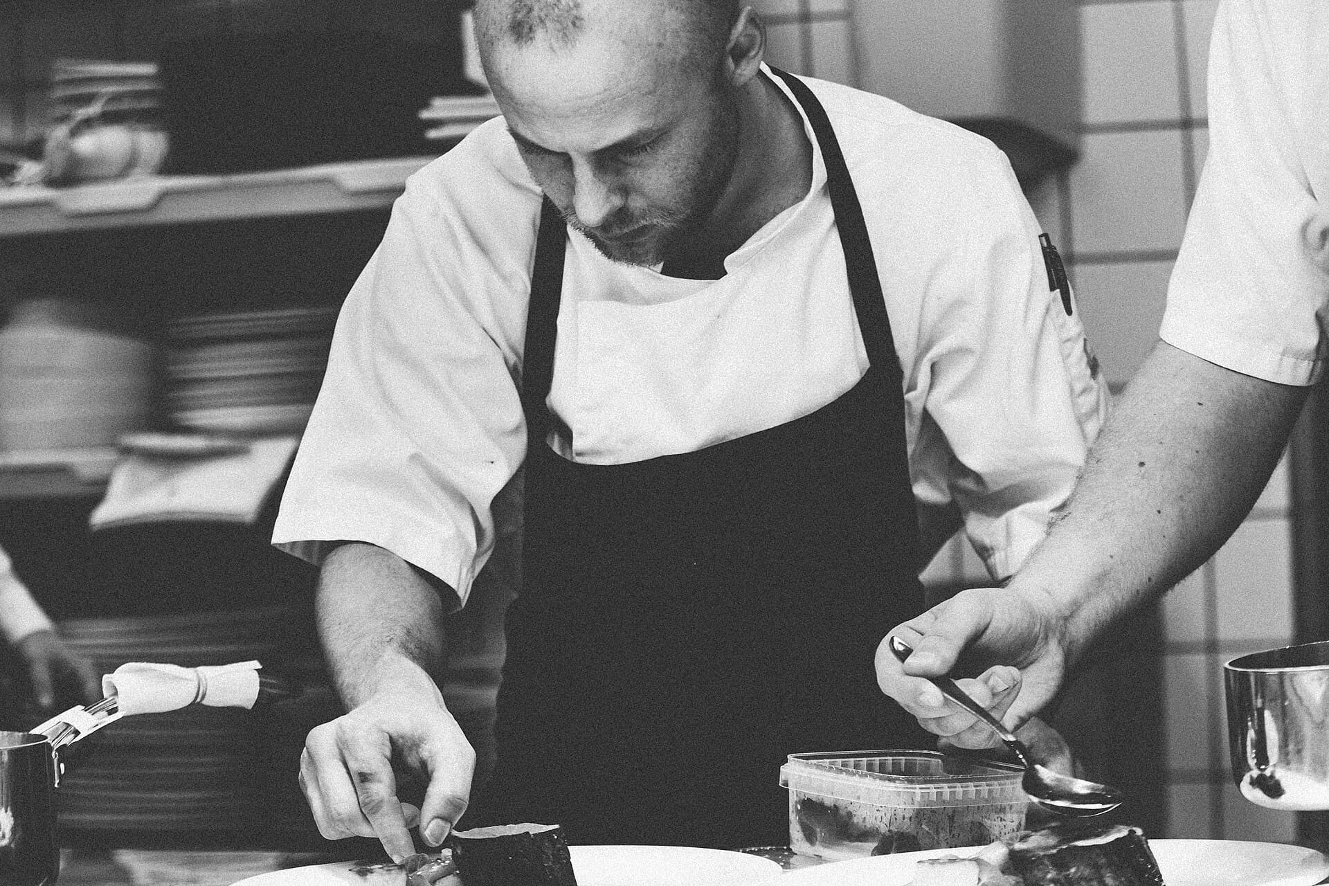 A chef in a white jacket and dark apron leans over the pass, plating by hand, in black and white.