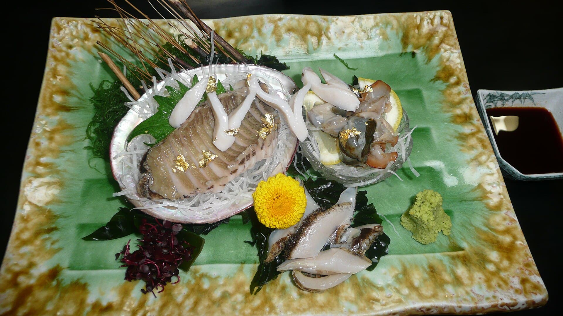 Steamed abalone with gold leaf and a yellow chrysanthemum on a green ceramic plate, beside a small dish of soy.
