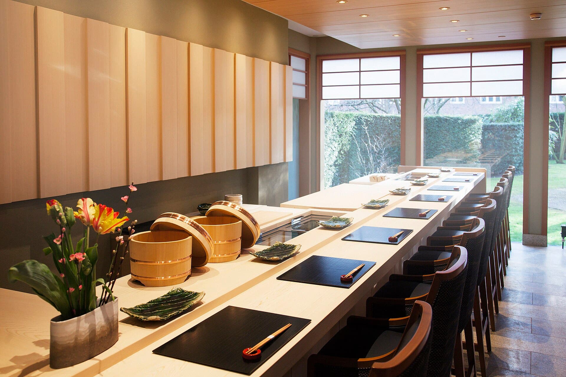 A long pale hinoki counter set for service, with lacquer trays, green ceramic plates and tall windows onto a garden.